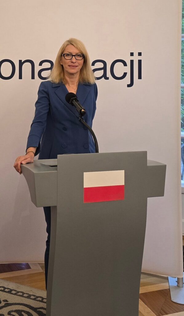 Paulina Malinowska-Kowalczyk stoi przy mównicy z biało-czerwoną flagą.